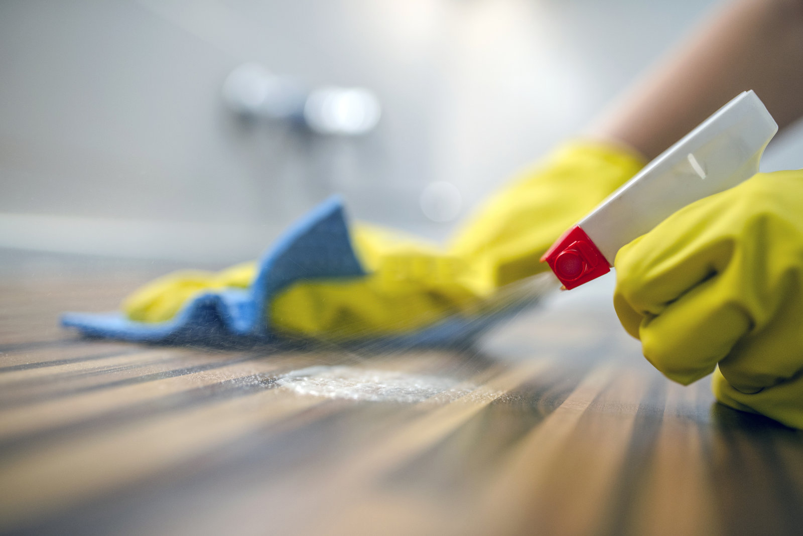 Fotografia ženy, ktorá čistí kuchynské skrinky špongiou a čističom v spreji.Žena používa sprejový čistič na drevený povrch.Slúžka utierajúci prach pomocou spreja a prachovky pri upratovaní domu v žltých ochranných rukaviciach, detail